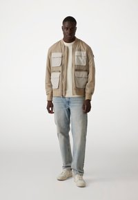 Veste bomber beige clair avec plusieurs patchs crème, poignets côtelés, une fermeture éclair à l'avant, portée sur une chemise claire et un jean bleu. Baskets blanches.