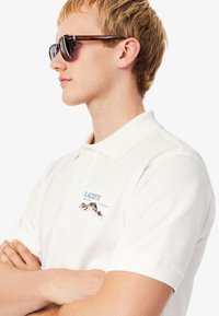 Vit pikétröja med texturerad tyg, med en broderad bergdesign och "LACOSTE" i blått. Bärs med mörka solglasögon.
