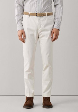 Man wearing white trousers, a striped button-up shirt, brown suede belt, and brown suede lace-up shoes standing against a plain background.