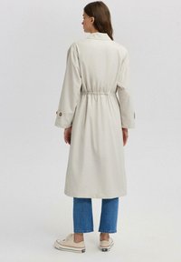 Trench coat bej deschis, cu talie strânsă, mâneci lungi și detalii cu butoane la mansete, asortat cu blugi blue cropped și tenisi albi.