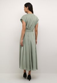 Long, flowing dress in sage green, with short sleeves, a belted waist, and a smooth, lightweight fabric. Elegant and minimalist design.