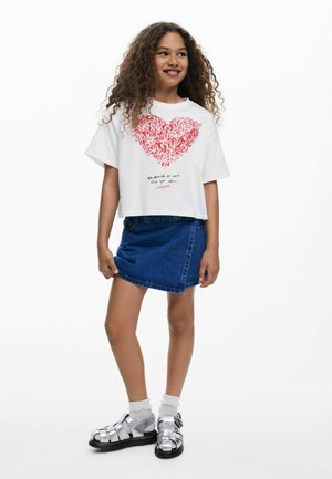 Camiseta blanca recortada con diseño de corazón rojo, falda de mezclilla y sandalias plateadas con tiras. Texto debajo del corazón dice "Siéntete orgulloso de amar quien eres."