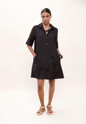 Mujer con vestido negro hasta la rodilla bordado con palmeras, joyas negras de cuentas y sandalias planas doradas, de pie con las manos en los bolsillos.