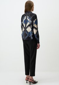 Langärmlige Bluse in Schwarz mit blauen und cremefarbenen Blumenmustern sowie Rautenmuster; kombiniert mit taillierten schwarzen Hosen und Absatzsandalen.