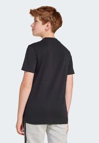 T-shirt noir à manches courtes en coton, avec un col rond classique et des lignes épurées, porté avec un pantalon gris. Vue de dos.