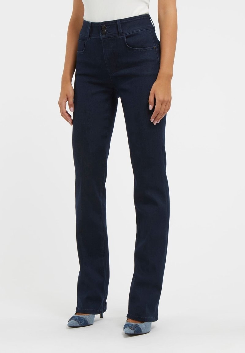 Femme portant un jean droit bleu foncé avec une taille à double bouton et des chaussures à talons bleues patchwork, debout devant un fond blanc uni.