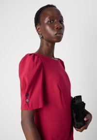 Burgunderkjole med pufferm, teksturert stoff og svarte knappeaksetter. Svart clutch med pelslignende tekstur holdt i hånden.