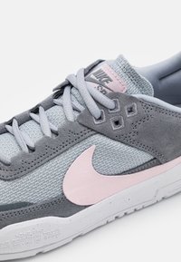 Nike tenisice s gornjim dijelom od sivog mrežastog materijala i ružičastim anilinskim detaljima, s teksturiranom sivom potplatom i svijetlosivim vezicama.