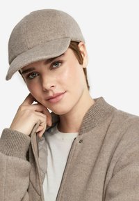 Grå ullkeps med rundad topp och styv skärm, bärs av en person i en ljusbrun bomberjacka över en vit skjorta.