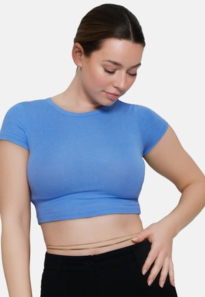 Joven mujer con top corto azul y pantalones negros, mirando hacia abajo con una mano en la cintura sobre un fondo liso.