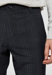 Tmavě šedé kalhoty na míru s jemným károvaným vzorem a hladkou texturou. Mají pas s knoflíkovým zapínáním a boční kapsy.