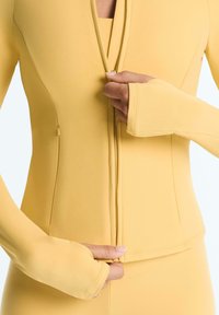 Mains fermant une veste jaune ajustée à manches longues avec des trous pour les pouces, montrant un gros plan de la zone du torse sur un fond uni.