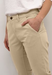 Beige Baumwollhose mit einer vorderen Tasche, Knopfverschluss und glatter Textur, die an einer Person gezeigt wird, die ihre Hand in die Tasche steckt.