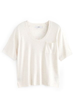 Woman wearing a cream short-sleeve top, beige mini skirt, and white ankle boots standing on wooden floor against pale green wall.