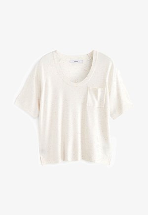 Kobieta ubrana w kremowy top z krótkim rękawem, beżową mini spódnicę i białe botki, stojąca na drewnianej podłodze na tle jasnozielonej ściany.