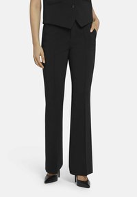 Zwarte broek van gladde stof, met een recht model, voorzakken en een lichte uitlopende zoom, gecombineerd met zwarte hakken.
