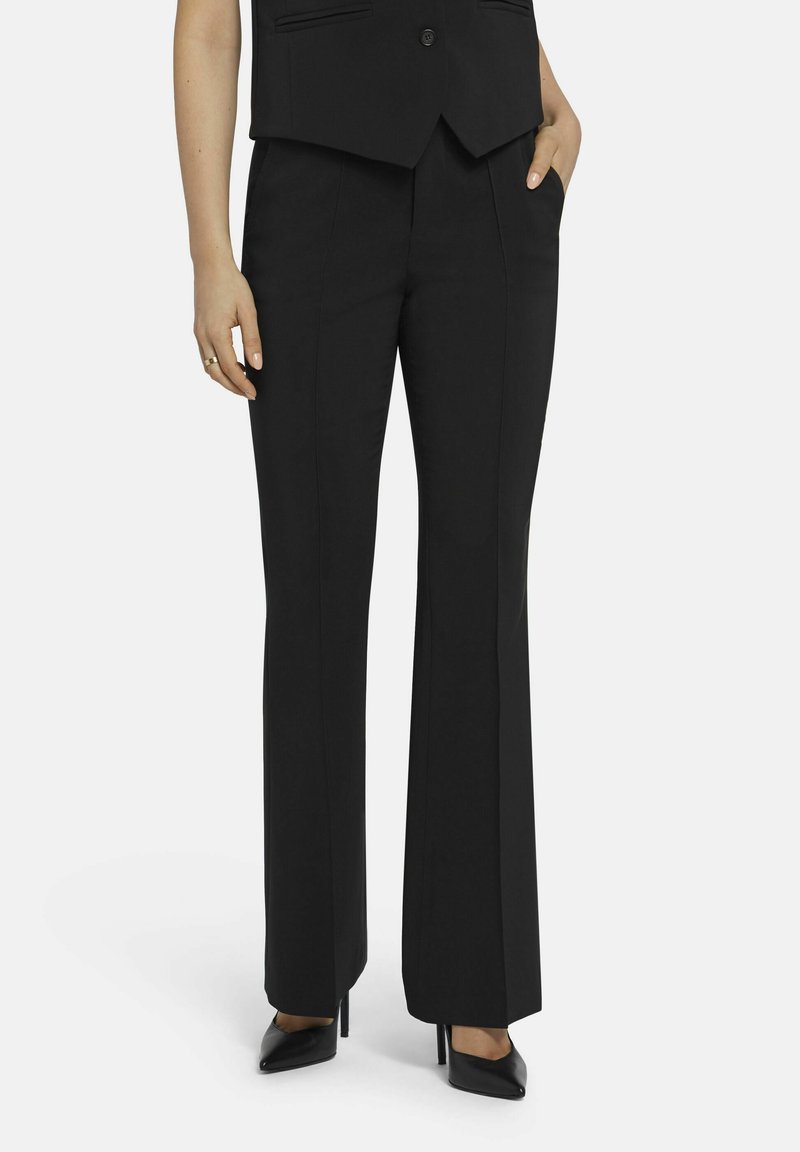 Zwarte broek van gladde stof, met een recht model, voorzakken en een lichte uitlopende zoom, gecombineerd met zwarte hakken.