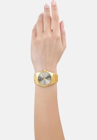 Gouden polshorloge met een zilvergrijze wijzerplaat, octagonale kast, gouden uurmarkeringen en een datumaanduiding. Metalen armband met een gepolijste afwerking.