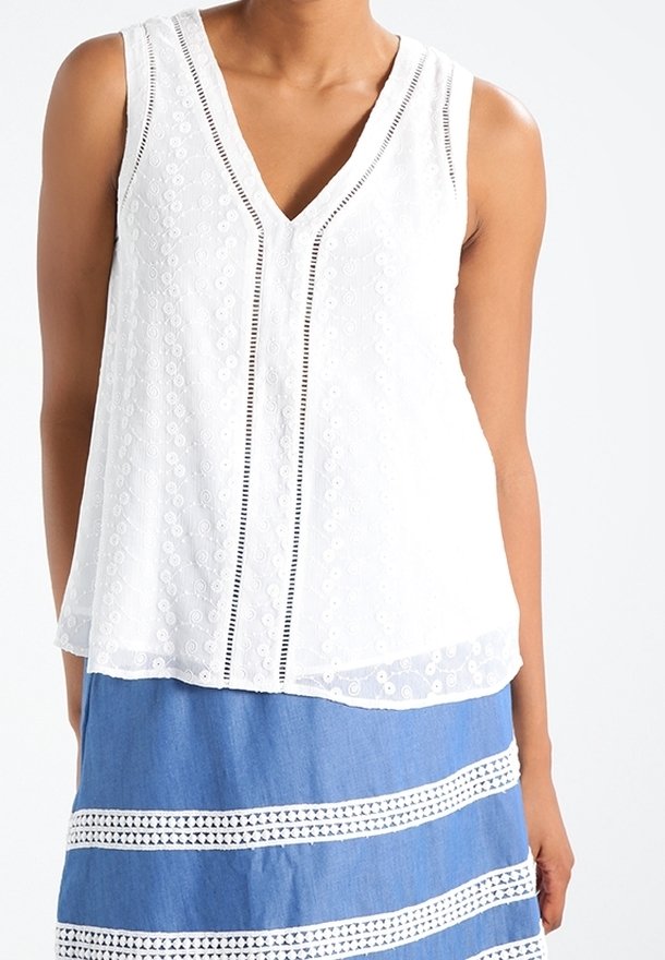 Haut sans manches blanc avec un décolleté en V, présentant des motifs floraux texturés et des rayures verticales noires sur le devant. Porté avec une jupe bleue.