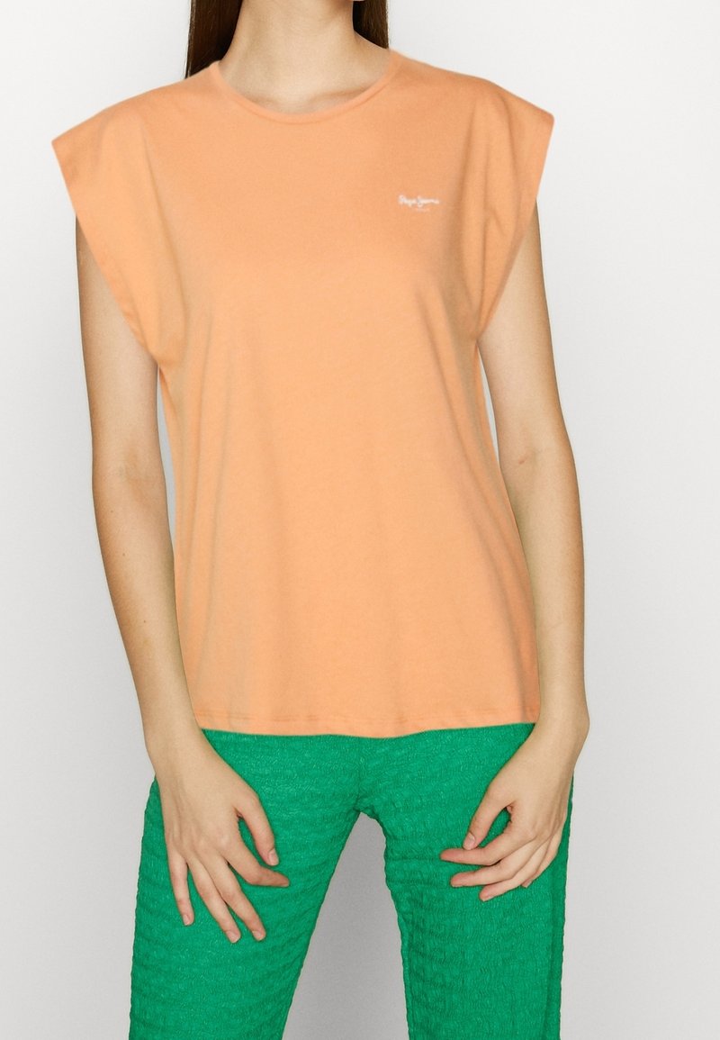 Tričko meruňkové barvy s krátkými, strukturovanými rukávy a kulatým výstřihem, vyrobené z jemné tkaniny. Obsahuje malé logo na hrudi.