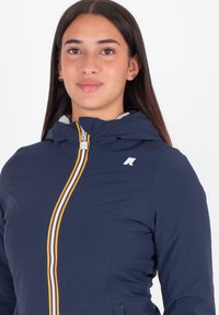 Giacca blu navy con zip e cappuccio, caratterizzata da una zip multicolore e un logo sul petto. Il materiale sembra liscio e leggero.