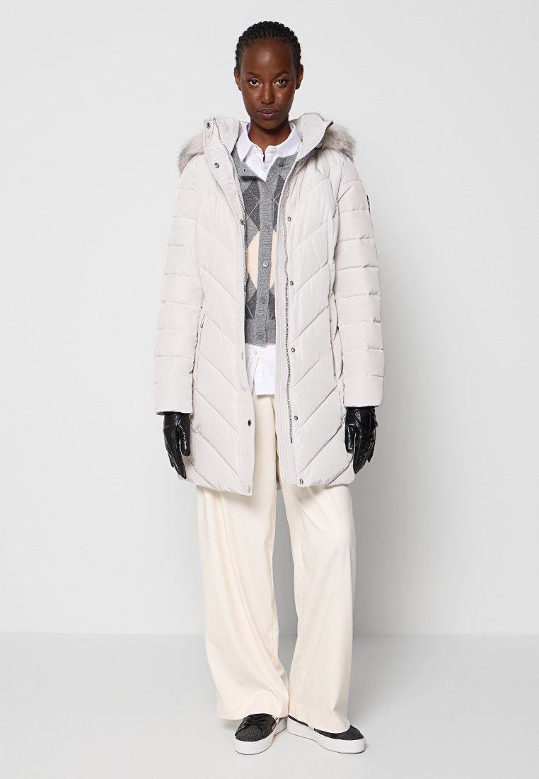 Beżowy płaszcz z ociepleniem, z kapturem i sztucznym futrem, z pikowanym wzorem, zapinany na zamek, noszony nad szarym swetrem w wzory i białymi spodniami.