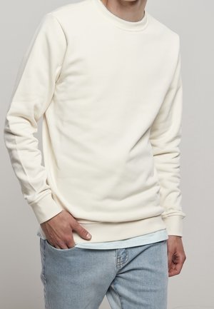 Felpa color crema realizzata in tessuto morbido. Collo a giro, maniche lunghe e polsini a costine. Indossata sopra una camicia azzurro chiara con jeans in denim.