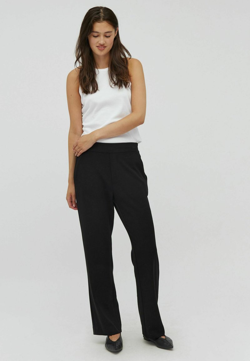 Zwarte rechte broek met een gladde textuur, gecombineerd met een witte mouwloze top. Het model poseert met één hand op haar heup.