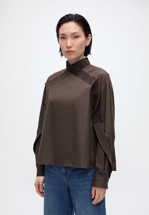 Femme aux cheveux courts et foncés portant un haut marron à col montant avec des détails plissés et un jean bleu, debout devant un fond clair uni.