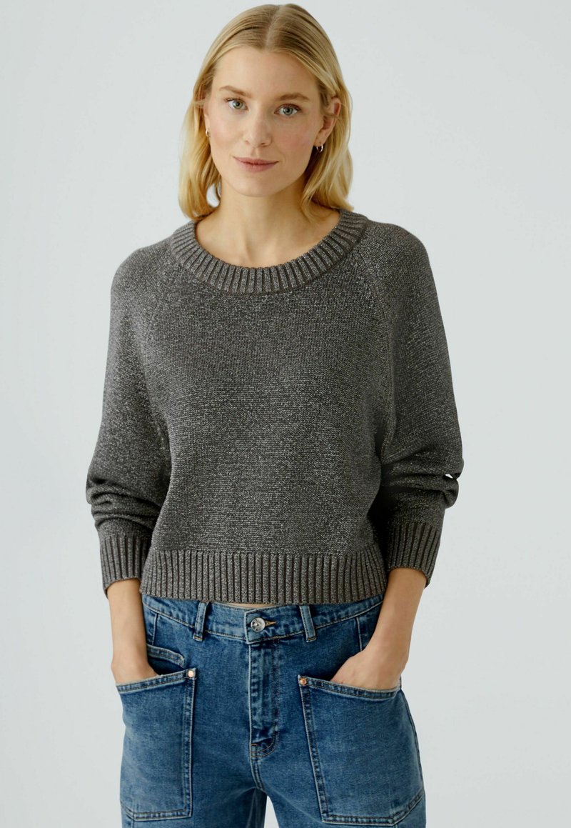 Grauer Strickpullover mit weitem Ripp-Ausschnitt und Bündchen, kombiniert mit lockeren blauen Jeans aus Denim, die große Vordertaschen haben.