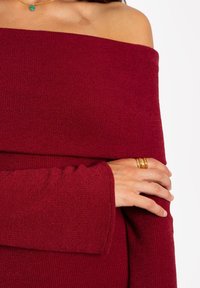 Pull en tricot bordeaux à épaule dénudée avec texture côtelée, doté de manches longues et d'une coupe décontractée, complété par une bague en or sur une main.