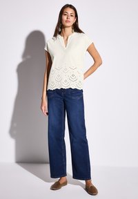 Lys beige kortærmet top med en bølget blondekant og øjelet detaljer, parret med mørkeblå bredbenede jeans og brune slip-on sko.