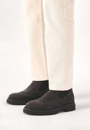 Dark brown suede ankle boots with brogue detailing, elastic side panels, and rubber soles, paired with light-colored, fitted pants.
