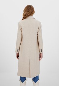 Cappotto lungo in misto lana beige con collo strutturato, fessura centrale e polsini a tre bottoni. Indossato sopra jeans blu e stivali chiari.
