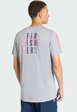 Mann trägt ein hellgraues kurzärmeliges Hemd mit dem Text "FINISHER 2025" und einer pinken Grafik auf dem Rücken, kombiniert mit dunkelblauen Shorts.