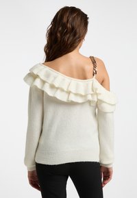 Creme kleurige off-shoulder trui met een volants halslijn, lange mouwen en een geribbelde zoom. Heeft een metallic kettingaccent op de schouderband.