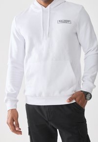 Mand iført hvid hættetrøje med teksten "BALANCE" og sorte cargobukser, hånd i lommen og sort armbåndsur mod ensfarvet baggrund.