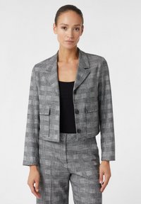 Blazer à carreaux gris coupé, avec des épaules structurées, des revers notch et deux poches avant. Porté sur un haut noir.