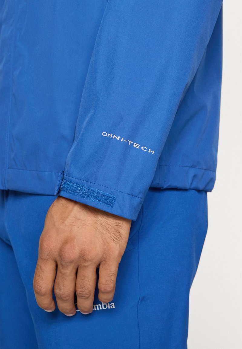 Niebieska wodoodporna kurtka z logo "OMNI-TECH" na rękawie, z regulacją mankietów na rzep i gładką, lekką teksturą.