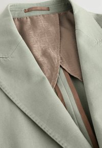 Lys grøn blazer med et glat, tekstureret stof. Indvendigt har den et mønstret beige foer og brune detaljer langs sømmene.