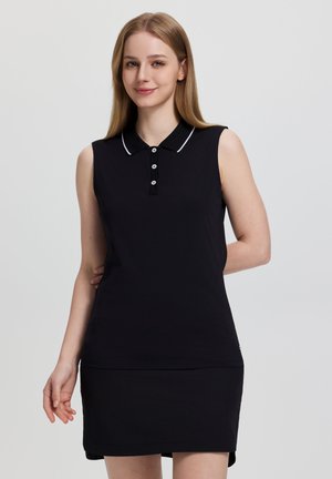 Nuori nainen pukeutuneena hihattomaan mustaan polomekkoon, jossa on valkoinen reunus kauluksessa, hymyilee yksinkertaisen vaalean taustan edessä.