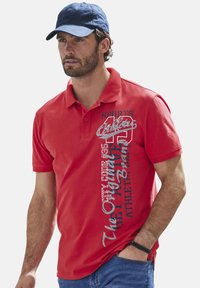 Rotes Poloshirt mit kurzen Ärmeln. Mit bedrucktem weißen und grauen Text. Aus weichem Baumwollmaterial. Kombiniert mit einer blauen Kappe.