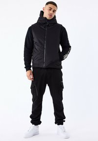 Sort polstret vest med høj krave og lynlås, parret med sorte cargo-bukser med lynlåslommer og hvide sneakers.