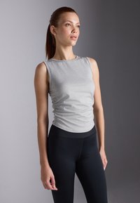 Model stands in a grey sleeveless top and black leggings, showcasing a relaxed pose against a neutral background, looking confidently to the side.