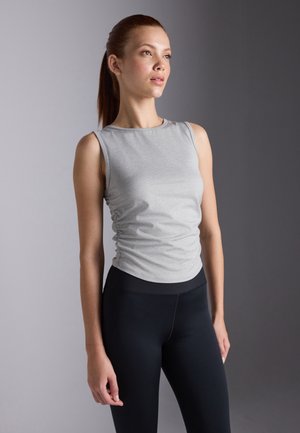 Modelen står i en grå ærmeløs top og sorte leggings, mens hun viser en afslappet positur mod en neutral baggrund, og ser selvsikkert til siden.