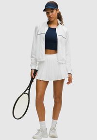 Woman holding a tennis racket wearing a navy crop top, white jacket, pleated white skirt, white socks, sneakers, and a navy visor.