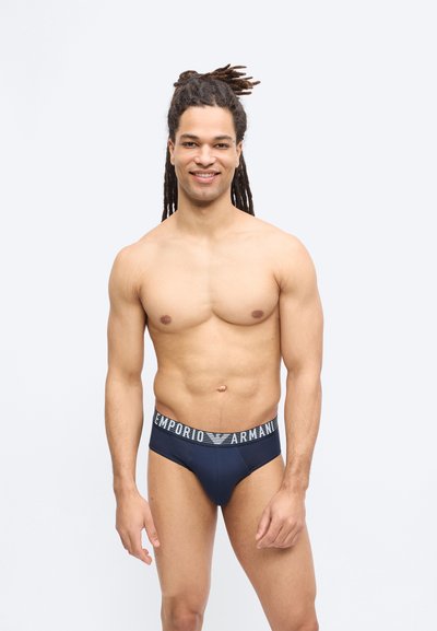 Χαμογελαστός άνδρας με dreadlocks που φοράει μπλε ναυτικό εσώρουχα Emporio Armani, στέκεται μπροστά σε ένα απλό ανοιχτό φόντο.