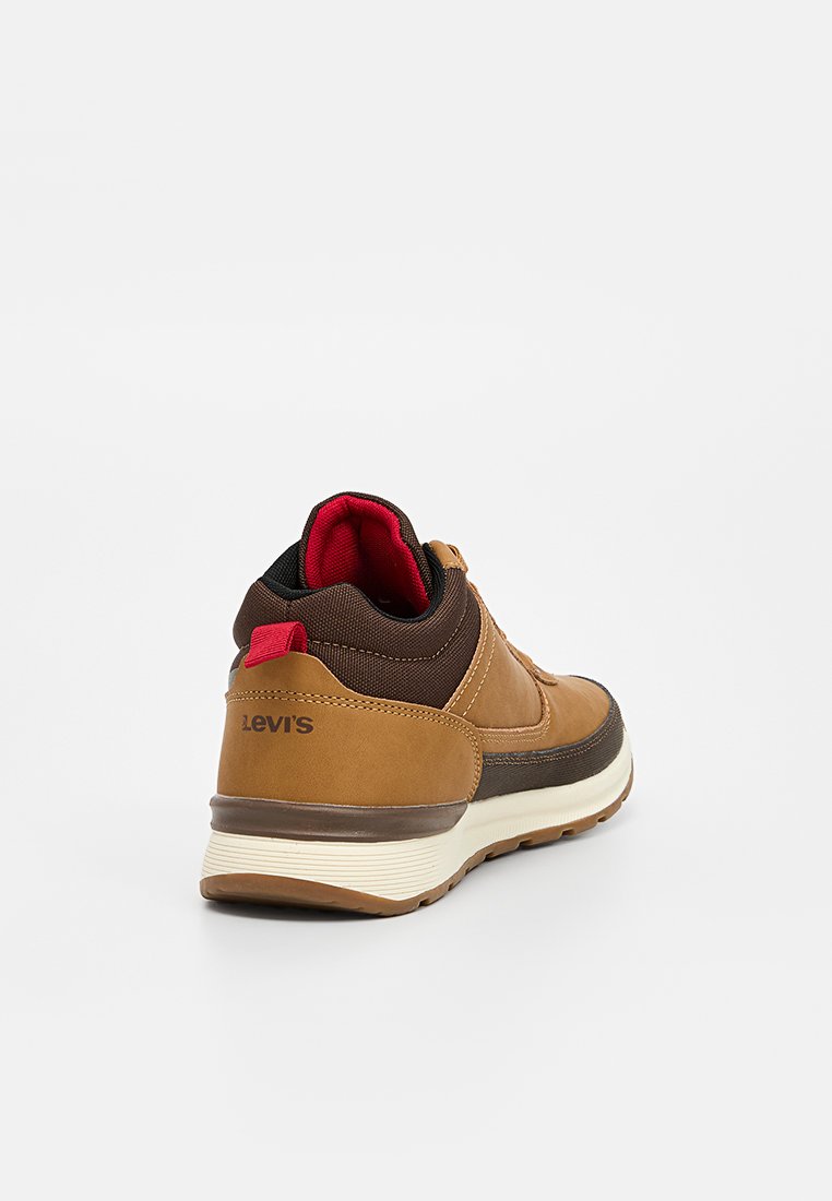 Sneaker in pelle marrone con colletto imbottito, dettagli rossi e suola bianca. Presenta una finitura testurizzata e un dettaglio del logo sul tallone.