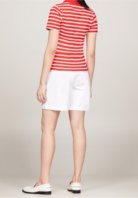 Polo a righe rosse e bianche con maniche corte, abbinato a pantaloni corti bianchi. Le scarpe sono mocassini bianchi. Sfondo neutro.