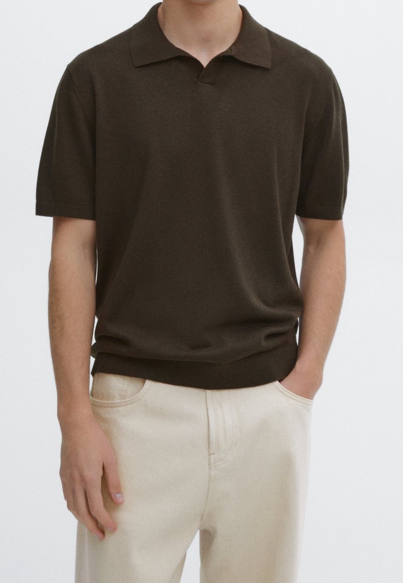 Homme portant un polo à manches courtes marron foncé, légèrement rentré dans un pantalon beige clair, une main dans la poche.